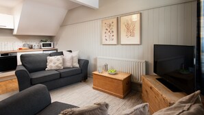 Ein Wohnzimmer mit grauem Sofa, einem Holztisch, einem Fernseher und einer Küchenzeile mit einer Mikrowelle.