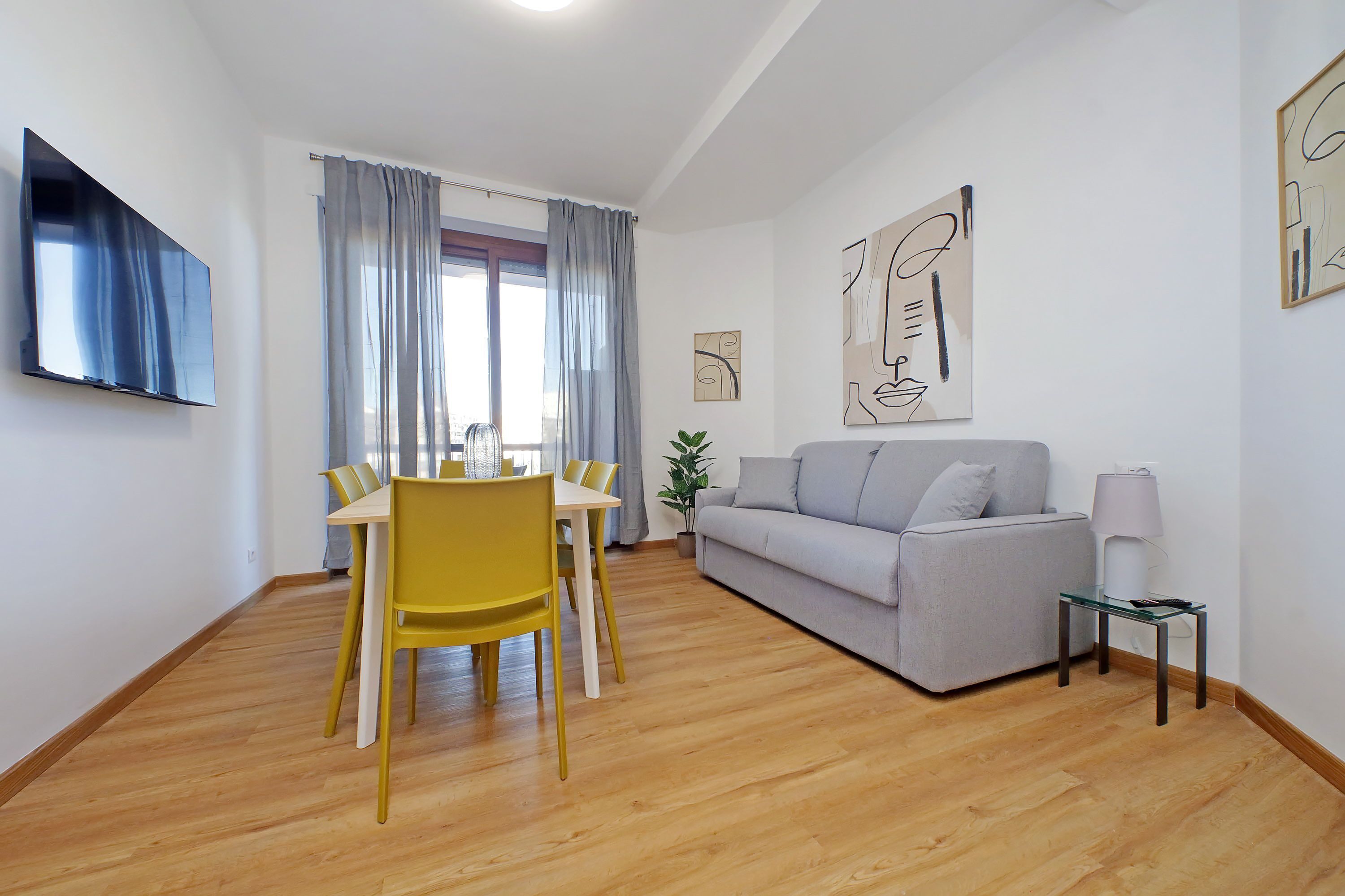 Un salon moderne comprenant un canapé, une table à manger avec des chaises, un téléviseur fixé au mur et des œuvres d’art accrochées aux murs.