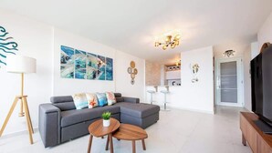 A modern living room with a grey sectional sofa, a wooden coffee table, and a TV on a wooden stand. There are abstract wall art pieces and a chandelier.