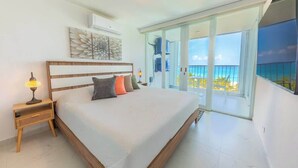 A bedroom with a large bed, a nightstand, a lamp, a ceiling light, a wall art piece, and a sliding glass door leading to a balcony with a view of the ocean.