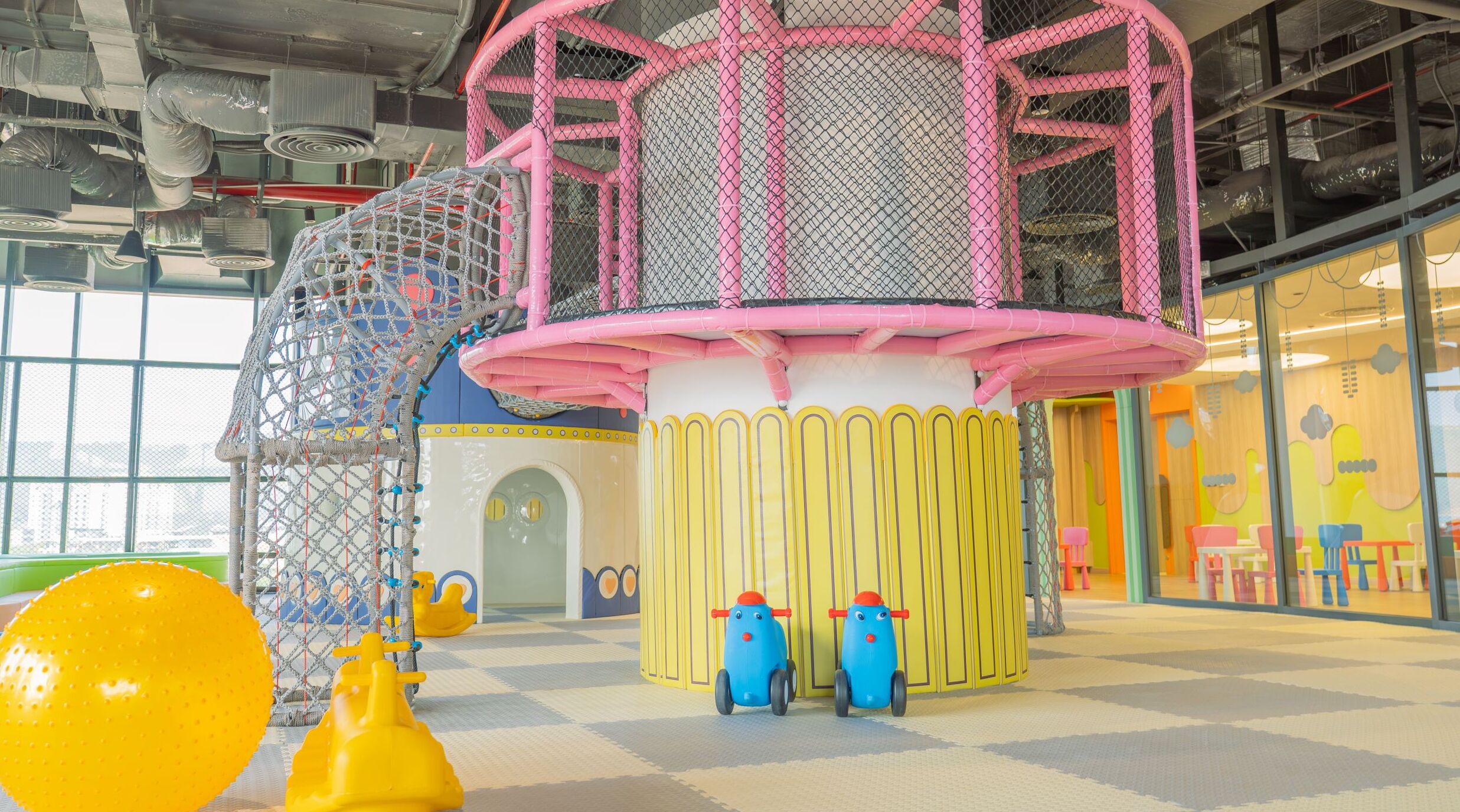 Un espace de jeux pour enfants avec une structure d’escalade en filet, une maison de jeu jaune et deux voitures jouets.