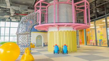 Un espace de jeux pour enfants avec une structure d’escalade en filet, une maison de jeu jaune et deux voitures jouets.
