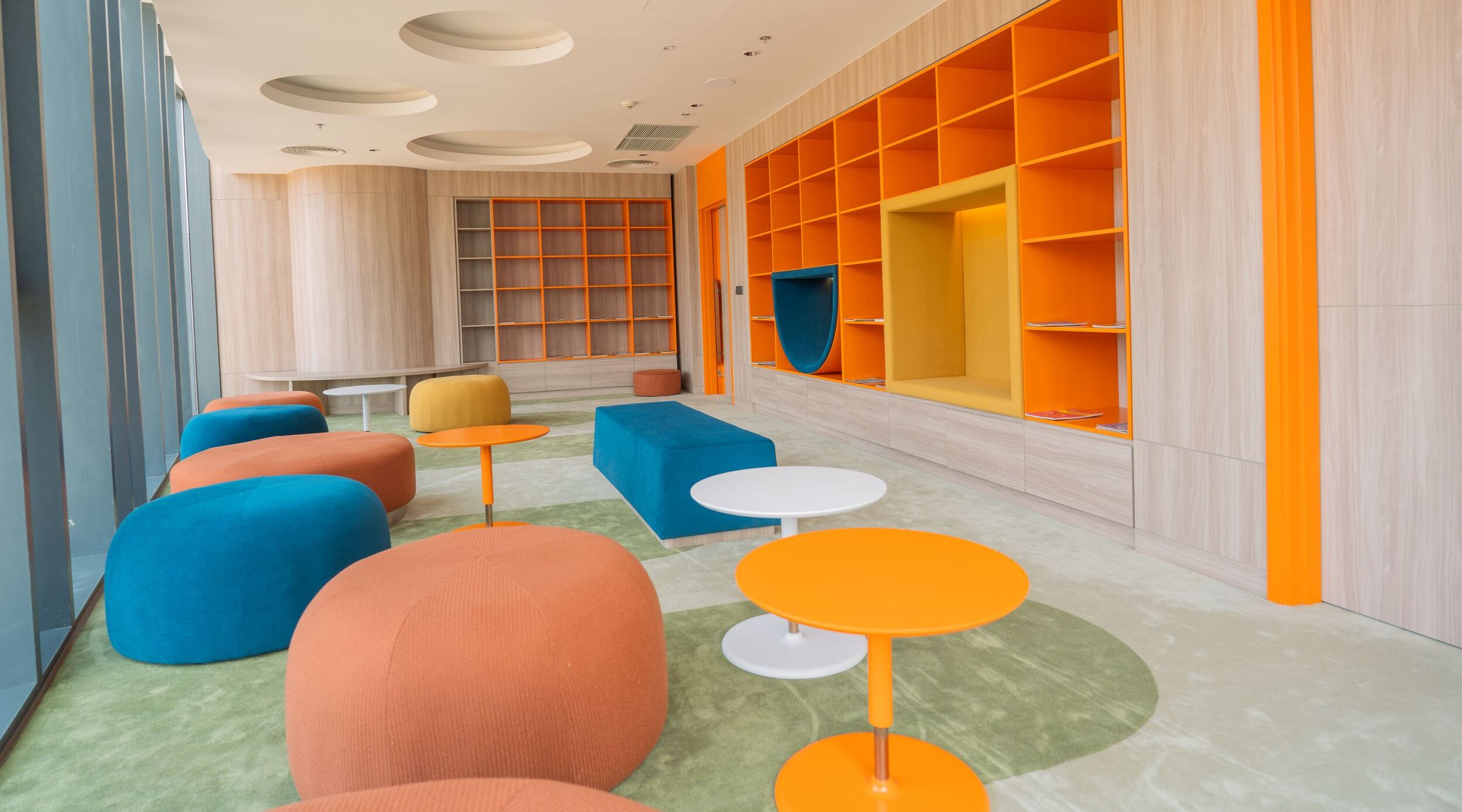 A modern lounge area with colorful seating, a large bookshelf, and a bright orange accent wall.