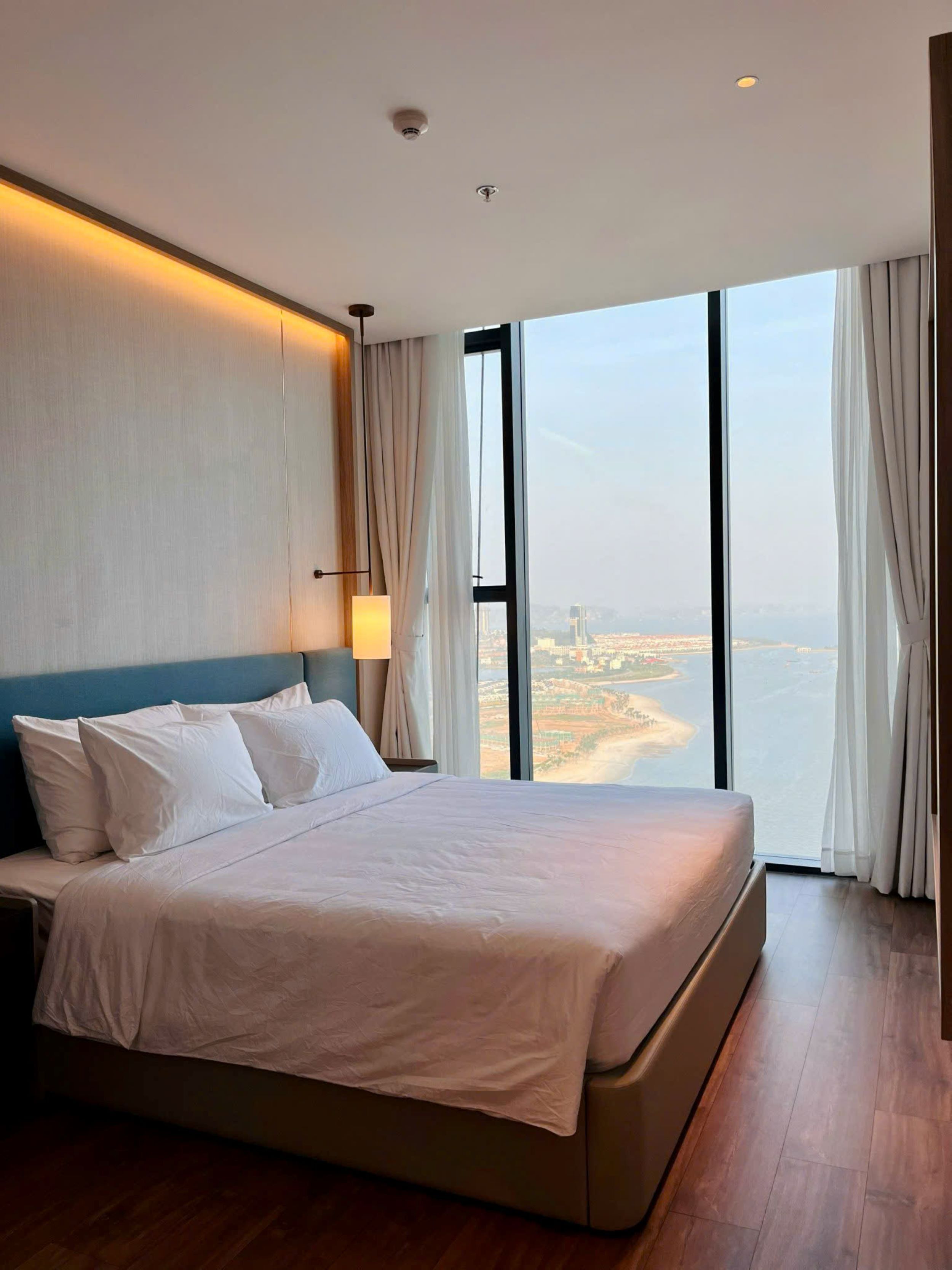 A modern hotel room with a large bed, a view of a coastal cityscape, and a large window with curtains.