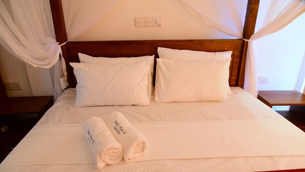 Una cama bien hecha con sábanas blancas, cabecera de madera y un par de toallas enrolladas con texto.