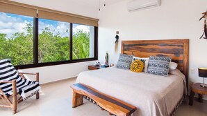 A bedroom with a large window, a bed with patterned pillows, a wooden bench, and a chair.