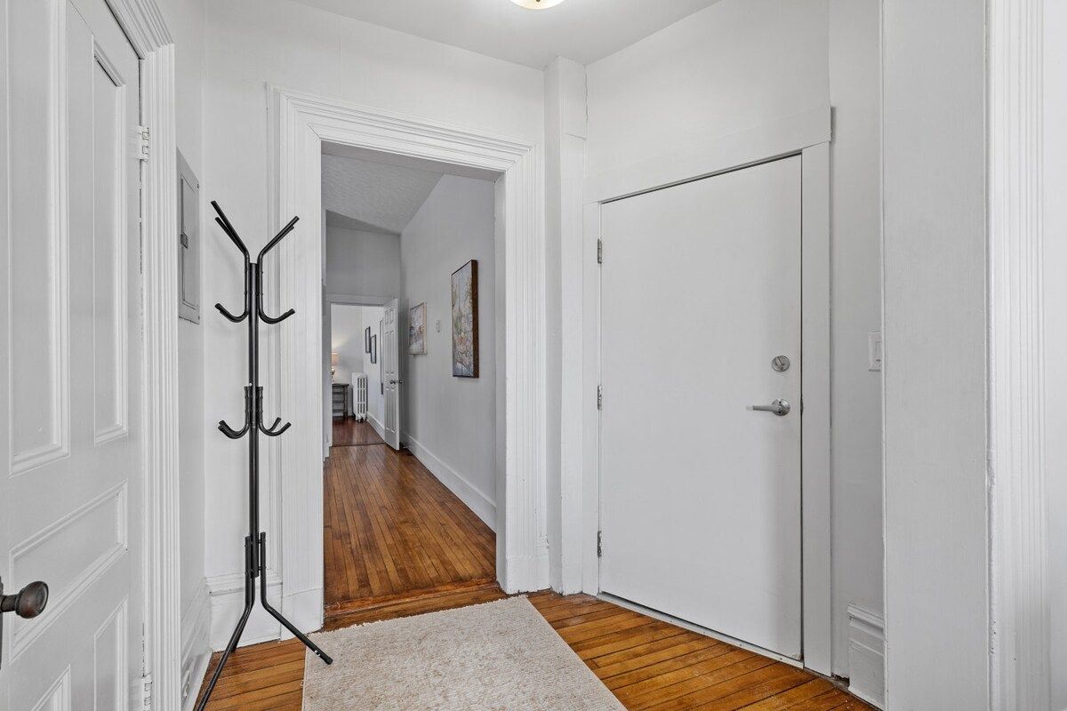 A hallway with white walls, wooden flooring, a coat rack, and a door.
