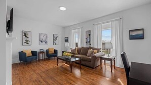 A living room with a brown sofa, two blue armchairs, a coffee table, and a TV on the left. - Downtown Urban Comfort Spacious 1 Bdrm Apartment (Charlottetown)