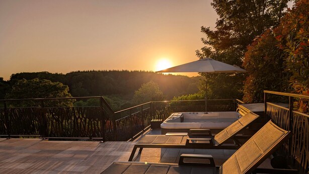 Ein Außenbereich mit Lounge-Möbeln und Whirlpool mit Blick auf eine bewaldete Landschaft bei Sonnenuntergang.