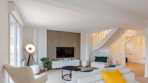 Ein modernes Wohnzimmer mit Flachbildfernseher, rundem Couchtisch und einer Couch mit Kissen. Eine Treppe führt zu einer weiteren Ebene.