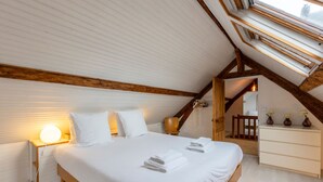 Ein Schlafzimmer mit schrägen Wänden, Holzbalken, einem Bett mit weißer Bettwäsche, einer Nachttischlampe und einer Kommode.