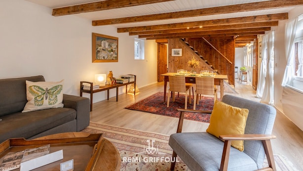 Ein geräumiges Wohnzimmer mit einer Couch, einem Sessel und einem Essbereich mit Tisch und Stühlen. Eine Treppe führt zu einem weiteren Raum.