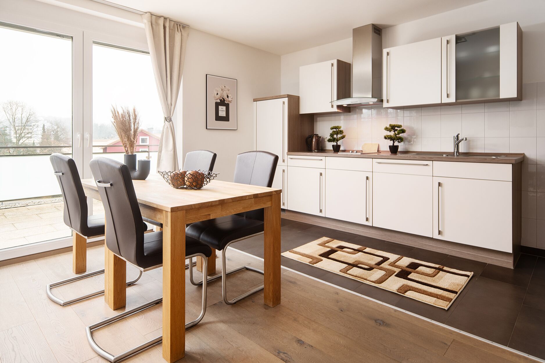 A modern kitchen with white cabinets, a wooden dining table, and a view through the window.