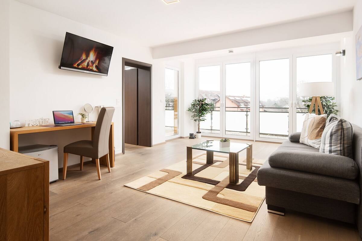 Un salon moderne comprenant un canapé, une table basse en verre, un bureau avec un ordinateur portable, un téléviseur et une grande fenêtre donnant sur un beau paysage.
