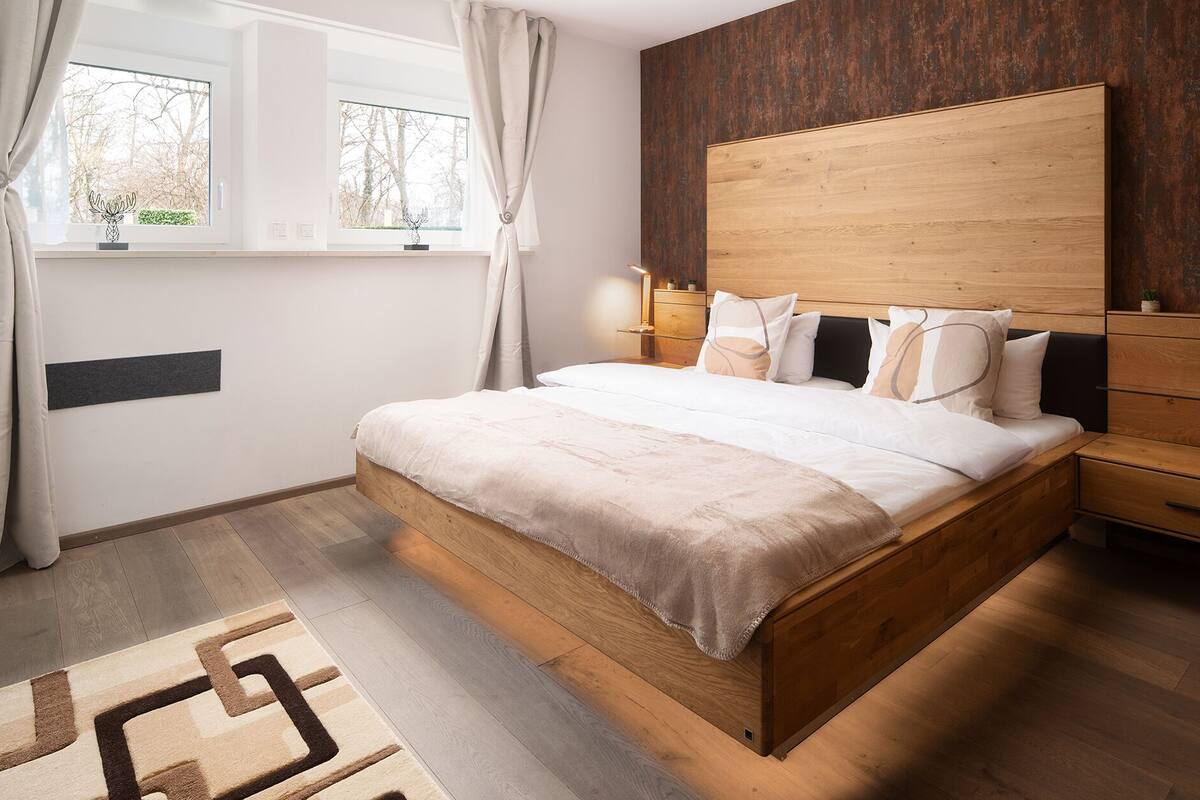 Une chambre à coucher avec un lit en bois, une table de chevet, une fenêtre avec des rideaux et une vue sur les arbres à l’extérieur.