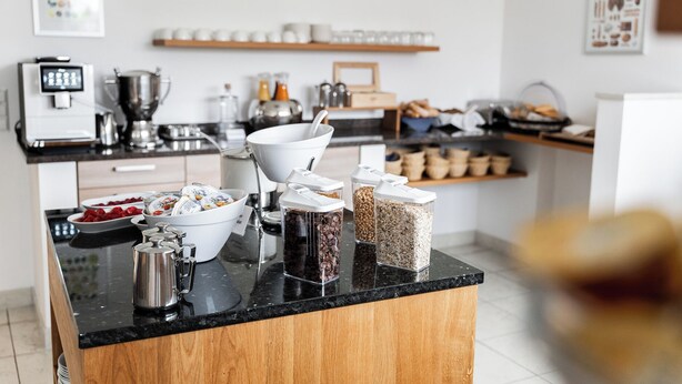 Een moderne keuken met een zwart aanrecht, houten kastjes en diverse etenswaren zoals yoghurt, granen en koffie.