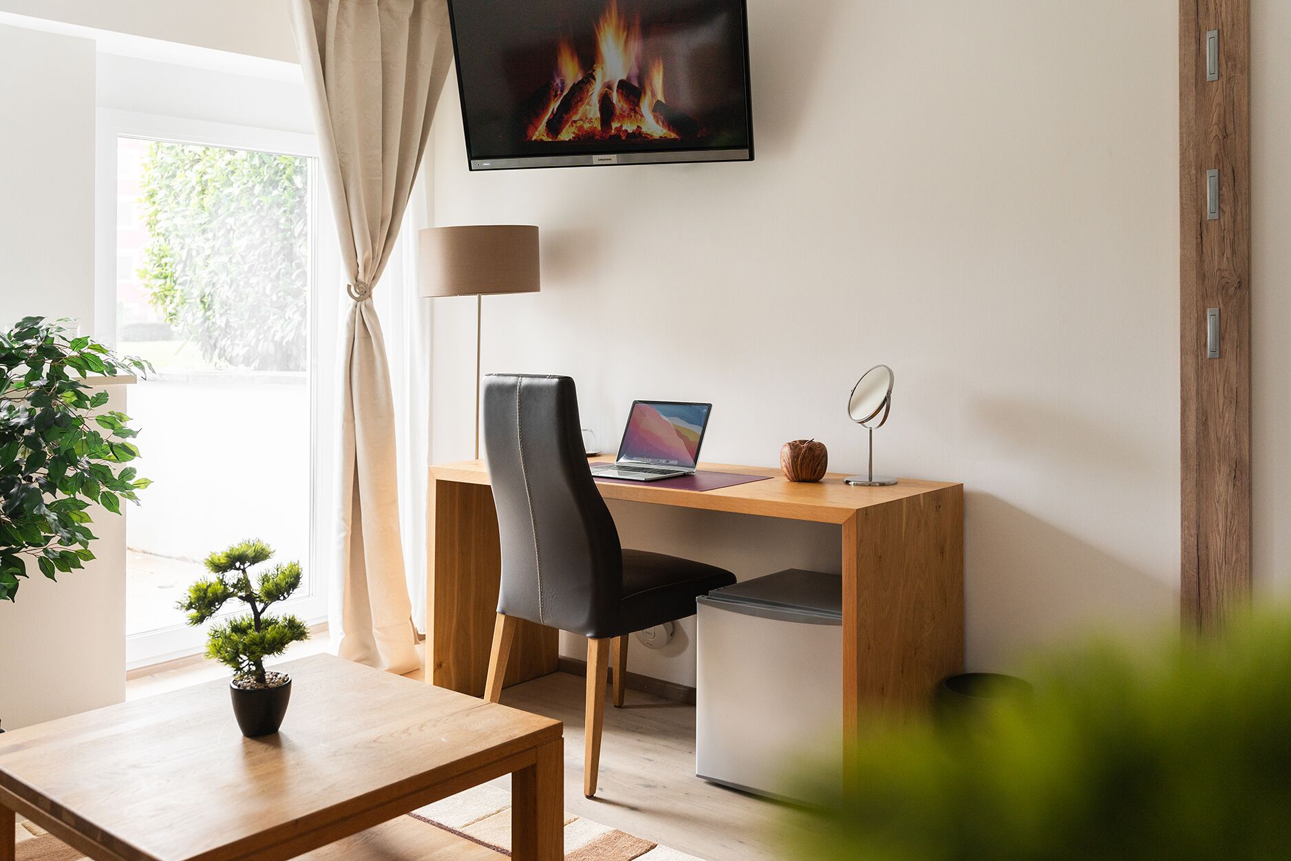 A modern room with a wooden desk, a laptop, a small mirror, and a television displaying a fire.