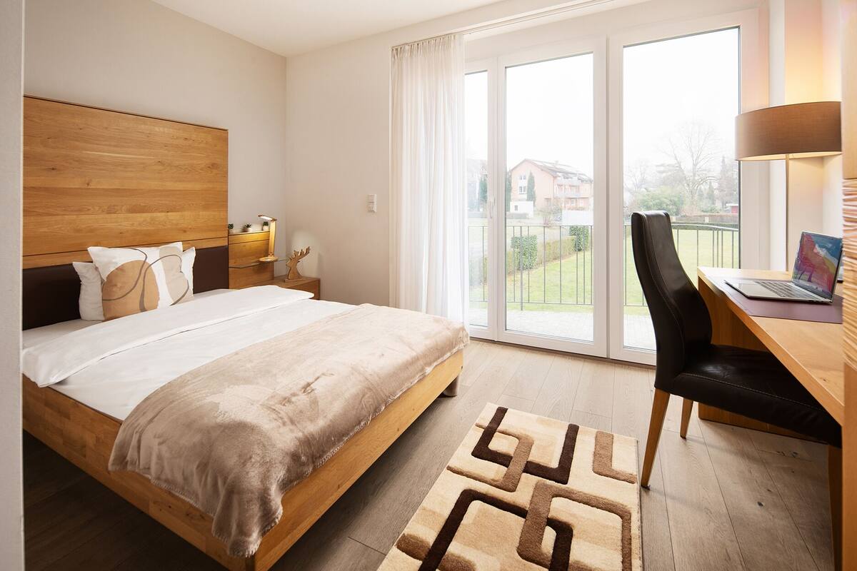 Une chambre à coucher avec un lit en bois, un bureau avec un ordinateur portable, et une vue sur un jardin grâce à une porte coulissante.