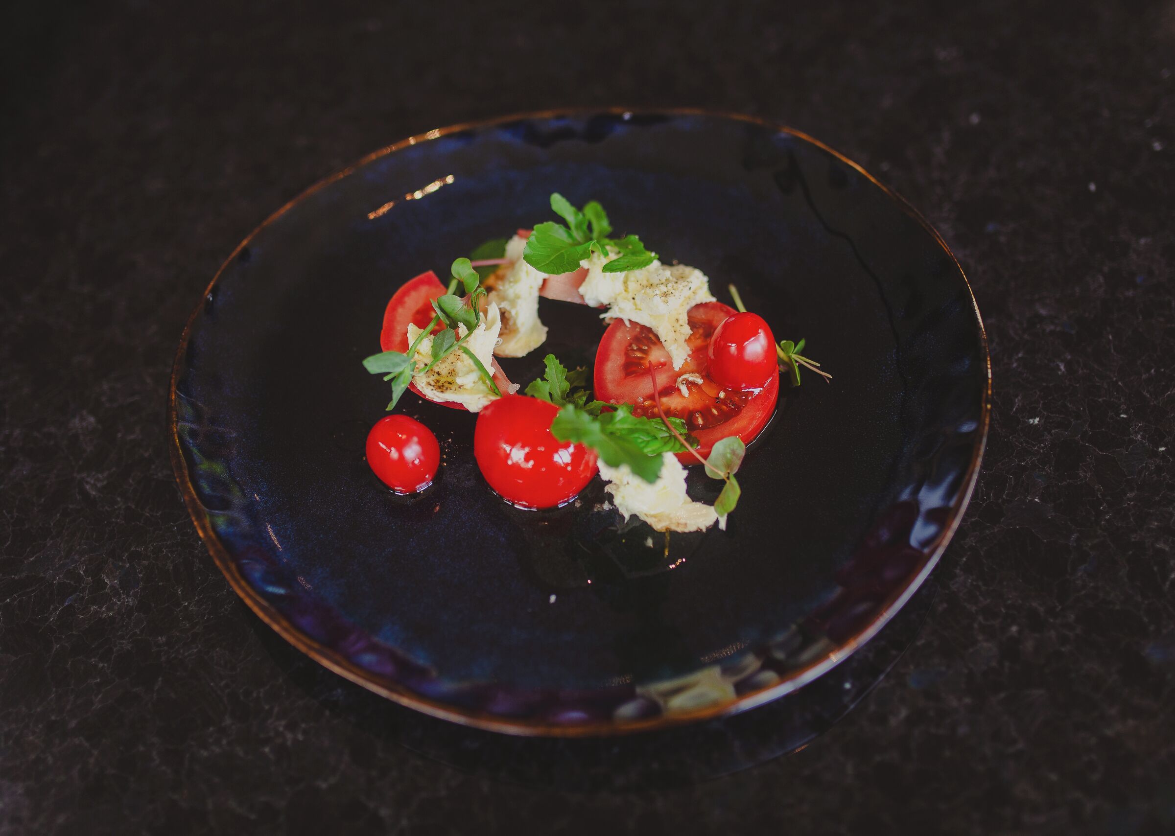 A dark plate with a food arrangement including tomatoes and greens.