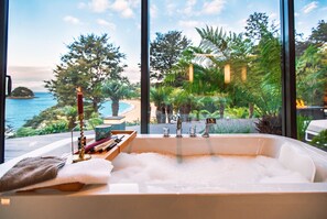 Een moderne badkamer met een groot bad, een houten dienblad met toiletartikelen, en uitzicht op een tropische tuin en de oceaan.
