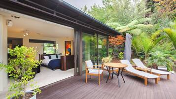 Terraza moderna al aire libre con mesa de madera y sillones de descanso, rodeada de abundante vegetación y una puerta de vidrio que da a un dormitorio con cama y un cuadro.