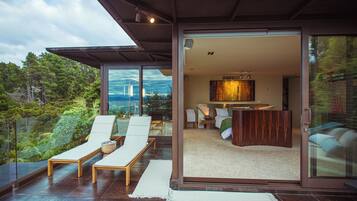 A modern interior with a glass door leading to a balcony with lounge chairs and a view of the forest.