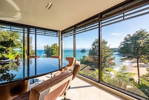 A modern dining area with a glass table and chairs, overlooking a scenic coastal landscape with trees and a view of the sea. - Exclusive Entire Lodge (Kaiteriteri)