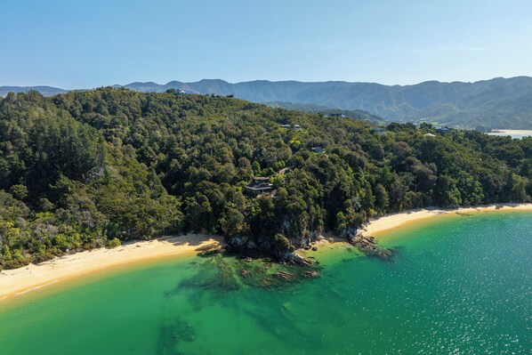 An aerial view of a coastal area with a beach, forested hills, and a small building on a cliff.