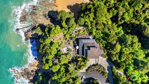 Vista aérea de uma propriedade costeira com uma casa cercada por vegetação densa, uma praia e um litoral rochoso.