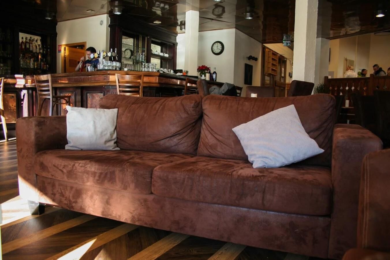 Un sofá de cuero marrón con dos almohadas blancas en un interior moderno con piso de madera y una zona de bar al fondo.