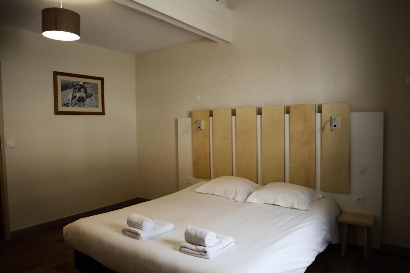 Habitación de hotel con una cama grande, cabecera de madera, dos almohadas, una mesita de noche, una pequeña mesa, un cuadro en la pared y una lámpara de techo.