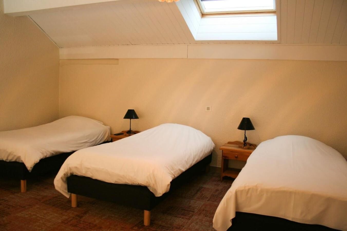 Habitación tipo dormitorio con tres camas individuales, cada una con una mesita de noche y lámpara. La habitación tiene un techo inclinado con tragaluz.