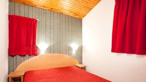 Een slaapkamer met een bed, een houten hoofdbord, rode gordijnen, een klein nachtkastje en een wandlamp.