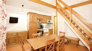 Een compacte keuken met houten kastjes, een eettafel en stoelen. Een kleine houten trap is rechts zichtbaar.