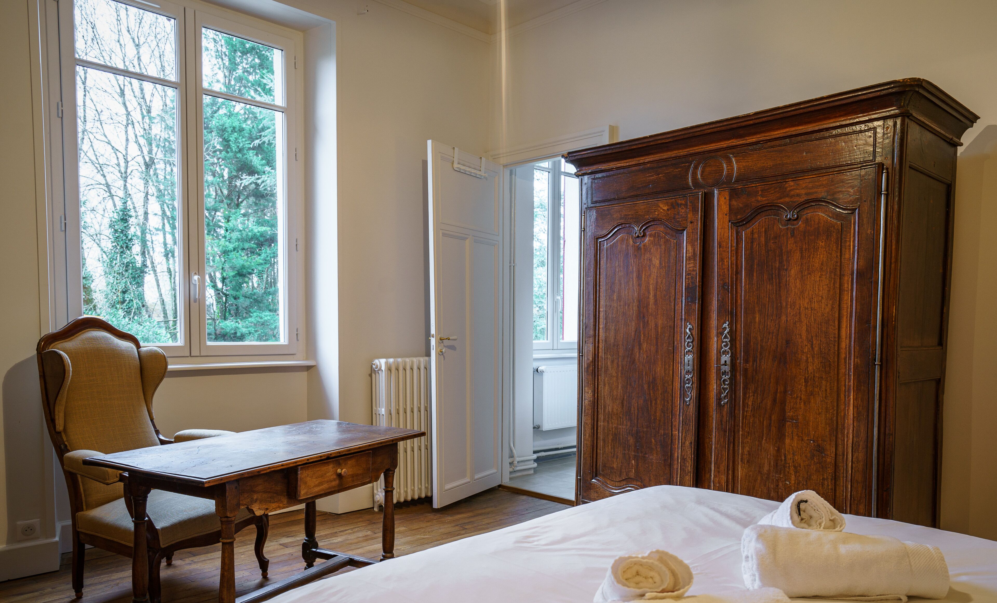 Une chambre à coucher avec une armoire en bois, une chaise en bois, une table en bois et une fenêtre donnant sur des arbres.