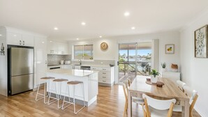 A modern kitchen with a large island, bar stools, and a dining area with a view of the marina. - Sunset Waters - Waterfront with Pool - Holiday Yamba (Yamba)