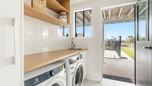 A compact laundry room with a washing machine, a sink, and wooden shelves. - Sunset Waters - Waterfront with Pool - Holiday Yamba (Yamba)
