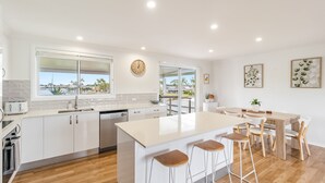 A modern kitchen with white cabinetry, a large island with bar stools, a view through the window, and a dining area with a wooden table and chairs. - Sunset Waters - Waterfront with Pool - Holiday Yamba (Yamba)