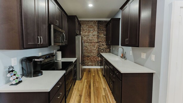 A modern kitchen with dark wooden cabinets, a brick accent wall, and a wooden floor. - Walk to Arch, Stadium & Dome! 2BR/2BA Downtown Gem ready for you and your family (St. Louis)