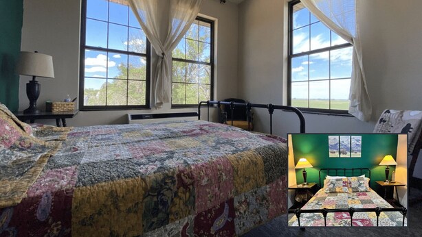 A room with a bed, two windows with curtains, a lamp, and a view of the outdoors. - Nome Schoolhouse Inn and Event Center (Nome)