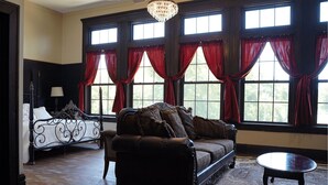 Una habitación con un sofá de cuero marrón oscuro, un sillón a juego y una pequeña mesa redonda. Amplias ventanas con cortinas rojas dejan entrar luz natural.