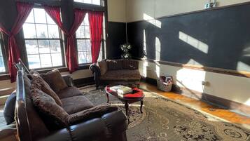Una sala de estar con un sofá de cuero marrón oscuro, un sillón estampado y una pequeña mesa roja. Hay un ventanal con cortinas rojas, una pizarra en la pared y un tapete con diseño en el suelo.