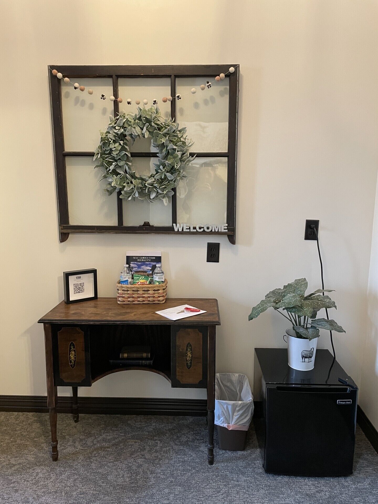 A desk with a welcome sign, a framed photo, a basket of bottled water, a potted plant, and a trash bin.