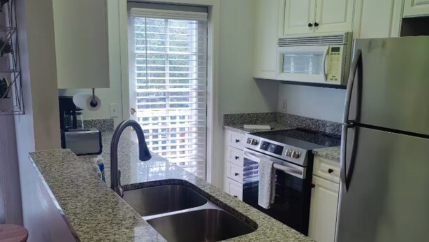 Una cocina con encimera de granito, pileta de acero inoxidable, muebles blancos, horno integrado y refrigerador.