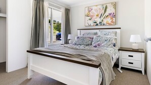 Une chambre à coucher avec un lit, des tables de chevet, une fenêtre avec des rideaux, un tableau et une vue sur l’extérieur.