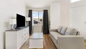 Un salon moderne avec un téléviseur à écran plat, un canapé blanc, une petite table blanche et une fenêtre avec des rideaux.