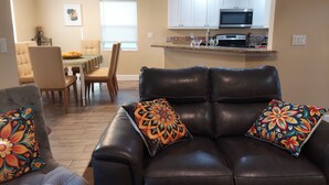 Una sala de estar con un sofá de cuero marrón, almohadones coloridos, un comedor con mesa y sillas, y una cocina con armarios blancos y un microondas.