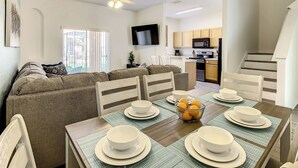A modern living room with a sofa, dining table set with bowls and plates, a chandelier, and a kitchen in the background. - Beautiful Townhome, Old Town (Kissimmee)