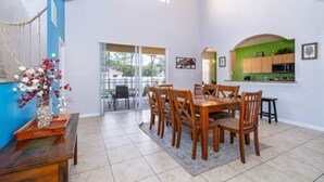 Uma sala de jantar com uma mesa e cadeiras de madeira, um vaso com flores, e vista para o exterior por uma porta de vidro.