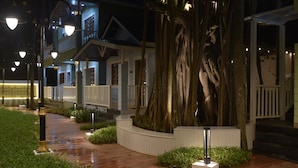 Un espacio exterior bien iluminado con un sendero de madera, un árbol grande y un edificio con balcón.